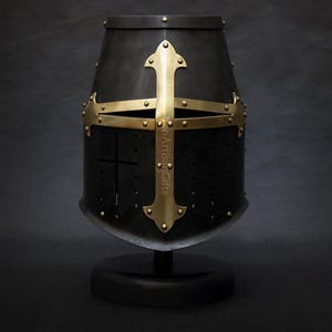 May include: A black medieval helmet with a gold cross design. The helmet is mounted on a black stand. The cross is riveted to the helmet. The helmet has small holes for ventilation.