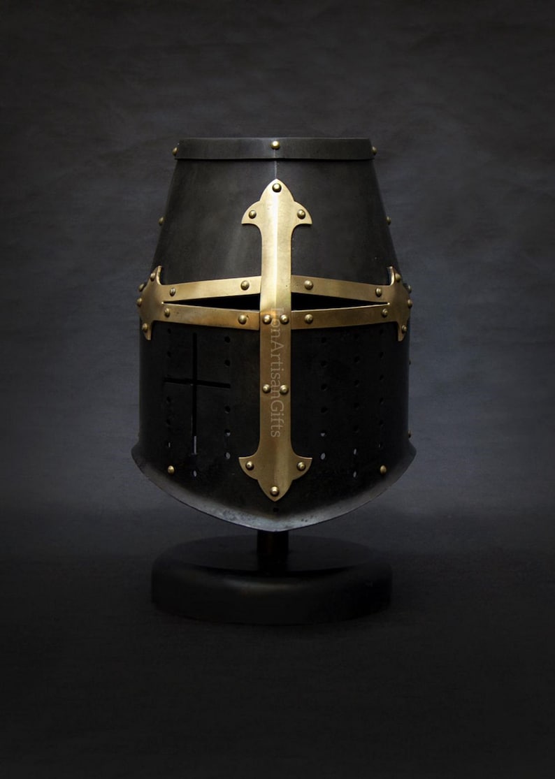 May include: A black medieval helmet with a gold cross design. The helmet is mounted on a black stand. The cross is riveted to the helmet. The helmet has small holes for ventilation.
