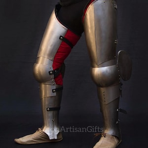 May include: A pair of steel leg armor, featuring articulated plates and knee protection. The armor is secured with leather straps and rivets, with red fabric visible between the metal plates. The image shows a side view of the armor.