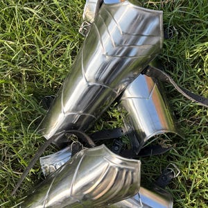 Könnte beinhalten: Poliertes Stahl-Armschutz-Set mit Lederriemen und Schnallen, das auf grünem Gras liegt. Die Rüstung hat eine reflektierende Oberfläche und ein segmentiertes Design, das mittelalterliche Themen suggeriert.