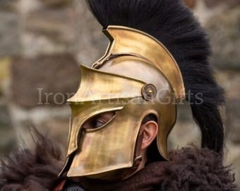 May include: A gold-colored Spartan helmet with a black plume. The helmet is part of a costume, worn with a brown fur collar and leather armor. The image is set against a blurred stone background.