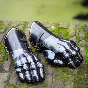 May include: Pair of black metal gauntlets with gold trim and rivets. The gauntlets are articulated at the knuckles and wrist, and are designed to protect the hands and forearms. The background is a brick wall with moss.