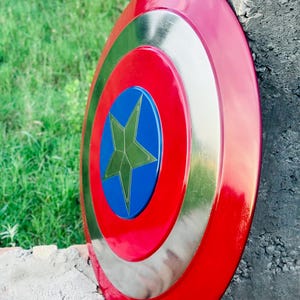 K&ouml;nnte beinhalten: Ein kreisf&ouml;rmiger Schild in Rot, Silber und Blau mit einem Sternemblem. Der Schild hat einen roten Au&szlig;enring, einen silbernen Mittelring und eine blaue Mitte mit einem goldenen Stern. Er lehnt an einer Betonstruktur.