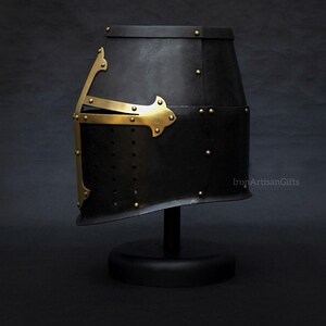 May include: Black medieval helmet with brass accents, rivets, and a cross design. The helmet is displayed on a black stand against a dark background. The product is from IronArtisanGifts.