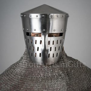 Op de afbeelding: Een glanzende zilveren ridderhelm met een conische top en een frontplaat met rechthoekige openingen. De helm is geklonken en wordt gedragen over een maliënkolder, met daaronder een rood kledingstuk.