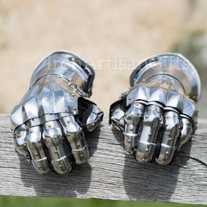May include: A pair of polished steel gauntlets, with articulated fingers and studded details. The medieval-style gloves are displayed on a weathered wooden surface, with a blurred natural background. The gauntlets are a shiny silver colour.