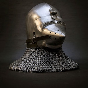 May include: A polished metal knight's helmet with a pointed visor and numerous ventilation holes. The helmet features a decorative element on top and a leather strap. A chainmail neck guard is attached to the helmet.
