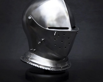 May include: A polished, silver-colored knight's helmet. The helmet features a visor with small holes, a neck guard, and rivets. The helmet is displayed on a black stand against a dark background.