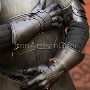 Puede incluir: Primer plano de guanteletes y armadura de metal negro. Los guanteletes están articulados con placas remachadas y segmentos de dedos. La armadura tiene un acabado metálico oscuro. El texto "Iron Artisan Gifts" es visible.