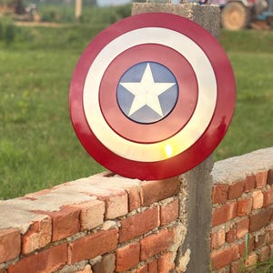K&ouml;nnte beinhalten: Runder Schild in Rot, Wei&szlig; und Blau mit einem wei&szlig;en Stern in der Mitte. Der Schild, mit gl&auml;nzender Oberfl&auml;che, erinnert an Captain America. Er ist an einem Betonpfosten neben einer Ziegelmauer befestigt.