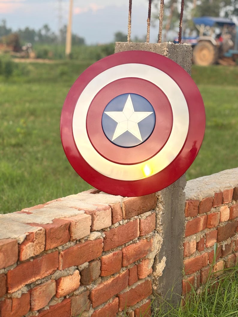 K&ouml;nnte beinhalten: Runder Schild in Rot, Wei&szlig; und Blau mit einem wei&szlig;en Stern in der Mitte. Der Schild, mit gl&auml;nzender Oberfl&auml;che, erinnert an Captain America. Er ist an einem Betonpfosten neben einer Ziegelmauer befestigt.