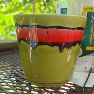 May include: A ceramic plant pot with a glossy finish. The pot is primarily olive green with a horizontal band of red and black. The pot has a slightly flared shape and is suitable for indoor or outdoor use.