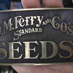 May include: A brass belt buckle with the text "D.M. Ferry and Co's Standard Seeds" in gold lettering on a black background.