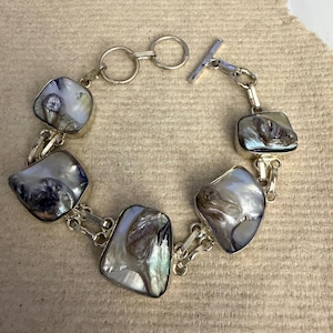 Op de afbeelding: Een zilveren armband met zeven onregelmatig gevormde, iriserende, witte en grijze stenen. De stenen zijn verbonden door zilveren schakels en hebben een tuimelsluiting. De armband wordt gepresenteerd op een neutraal gekleurd oppervlak.