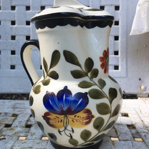 Może przedstawiać: Biały dzbanek ceramiczny z pokrywką, ozdobiony ręcznie malowanymi wzorami kwiatowymi w odcieniach niebieskiego, żółtego, zielonego i czerwonego. Dzbanek ma czarną obwódkę i czarną podstawę.