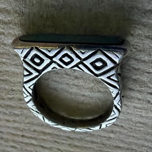 May include: A silver ring with a rectangular, dark green stone. The ring has a geometric pattern on the band and a gold-colored border around the stone. The ring is likely made of metal and is designed for wear on a finger.