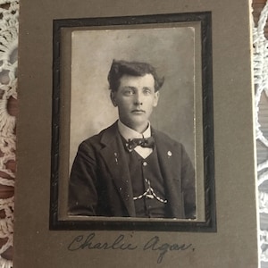 Könnte beinhalten: Schwarz-Weiß-Porträt eines Mannes in Anzug, Fliege und Weste. Das Foto ist in einem dunklen Rahmen mit dem handschriftlichen Namen "Charlie Agau" unten. Das Foto liegt auf einer weißen Spitzendecke.