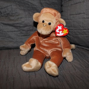 May include: A brown plush monkey wearing a brown beanie with a red Ty tag attached to the beanie. The monkey is sitting on a gray textured surface.