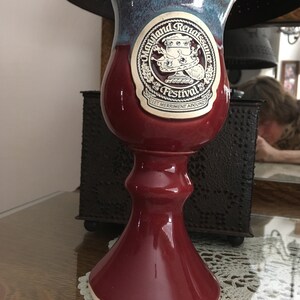 Puede incluir: Un cáliz de cerámica burdeos y azul-púrpura. El cáliz tiene una pegatina con las palabras "Maryland Renaissance Festival" y una imagen de dibujos animados. El cáliz está sobre una mesa con un tapete de encaje. El cáliz mide unos 18 cm de altura.
