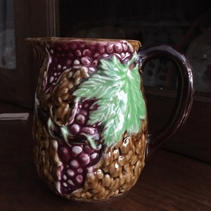 Può includere: Un'anfora in ceramica marrone con un motivo a vite. L'anfora ha un manico marrone ed è decorata con foglie verdi e uva viola.