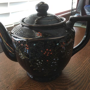 Puede incluir: Tetera negra con ribetes dorados y un patrón floral. La tetera tiene una tapa y un asa. La tetera está decorada con flores rojas, azules y blancas.