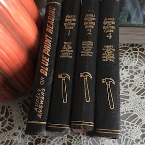 Könnte beinhalten: Ein Satz von vier schwarzen, ledergebundenen Büchern mit dem Titel "Audels Carpenters and Builders Guide" mit goldenen Lettern und Abbildungen von Hämmern. Die Bücher sind mit 1, 2, 4 und "Answers on Blue Print Reading" nummeriert.