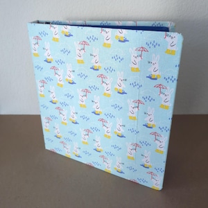 May include: A light blue binder featuring a pattern of white rabbits in yellow wellington boots, holding red umbrellas, and set against a rainy backdrop. The raindrops are blue.