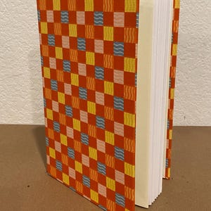 May include: A hardcover journal with a vibrant orange cover featuring a repeating pattern of squares in yellow, blue, and pink. The journal's pages are visible, and the spine is a light beige color.
