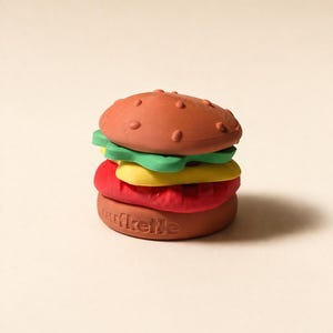 Peut inclure: Un jouet en forme de hamburger avec un pain brun, de la laitue verte, du fromage jaune et une galette rouge. Le pain du bas porte l'inscription "urfikette". Un jouet coloré et amusant.