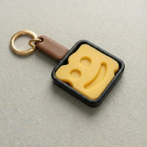 May include: A novelty keychain featuring a yellow cheese slice with a smiling face, set in a black square pan. The keychain has a brown leather handle and a gold-colored ring.
