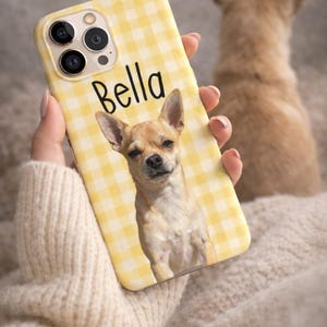 May include: A yellow phone case with a gingham pattern and the name "Bella" printed on it. The case features a photo of a small, light brown dog. The dog is a Chihuahua. The phone case is held by a person wearing a cream-colored sweater.