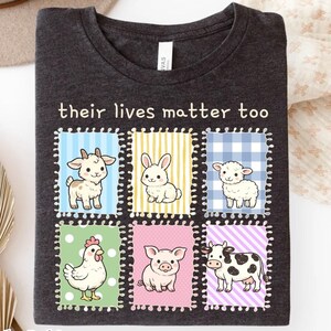 Op de afbeelding: Een donkergrijs gemêleerd t-shirt met de tekst "their lives matter too" boven zes kleurrijke rechthoekige vlakken. Elk vlak bevat een cartoonillustratie van een boerderijdier: een geit, konijn, schaap, kip, varken en een koe.