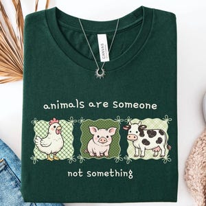 Op de afbeelding: Een donkergroen t-shirt met een afbeelding van een kip, een varken en een koe in individuele groene ruitjesvierkanten. De tekst "animals are Someone" staat boven de dieren en "not something" staat eronder.