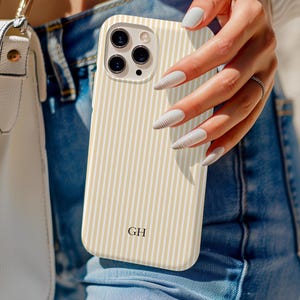 May include: A cream-colored phone case with vertical beige stripes. The case has a camera cutout and the initials "GH" at the bottom. The phone case is held in front of a person wearing blue jeans.