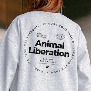 Op de afbeelding: Een lichtgrijs sweatshirt met ronde hals en zwarte tekst op de achterkant. Het ontwerp bevat centraal de woorden Animal Liberation, omringd door een cirkelvormige rand met de tekst Choose Compassion, Speaking For Them, Every Life Deserves Freedom.