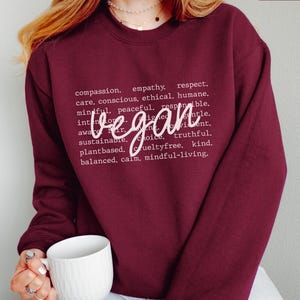 Puede incluir: Sudadera granate con la palabra "vegan" en cursiva. La sudadera también presenta una lista de palabras como "compasión", "empatía" y "sostenible" en fuente blanca. Una taza blanca se sostiene en la mano de la persona.