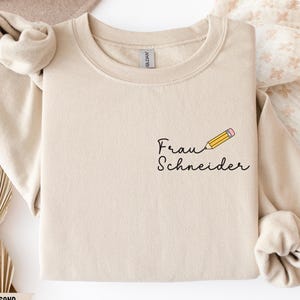 May include: A sand-coloured sweatshirt featuring the text "Frau Schneider" and a pencil graphic. The pencil is yellow with a pink eraser. The sweatshirt has a crew neck and long sleeves.