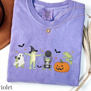 May include: A violet short-sleeved t-shirt with a Comfort Colors label at the neck. The graphic features four frogs in Halloween costumes: one as a ghost, one as a witch, one as a skeleton, and one jumping near a jack-o'-lantern, with bats and stars.