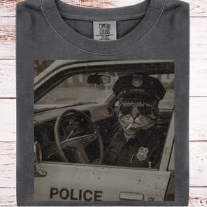 May include: Dark gray t-shirt featuring a sepia-toned image of a cat in a police uniform, wearing sunglasses and smoking a cigarette while sitting in a police car. The word "POLICE" is visible on the car door.