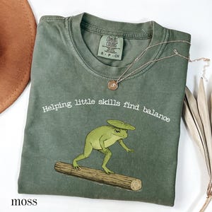 May include: Moss green t-shirt with a frog graphic balancing on a log, and the text "Helping little skills find balance." A gold necklace and a brown hat are also visible.