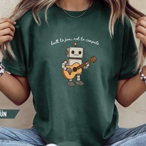 Può includere: T-shirt verde scuro con un robot che suona la chitarra. Il robot è beige e grigio, con la scritta "built to jam, not to compute" sopra. La maglietta è di colore uniforme.