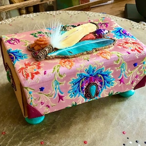 Könnte beinhalten: Eine dekorative rechteckige Box mit einem rosa Blumenmuster, verziert mit Federn und Perlen. Die Box hat türkisfarbene Füße und burgunderfarbene Eckakzente. Die Oberseite zeigt eine gelbe und türkisfarbene Feder.