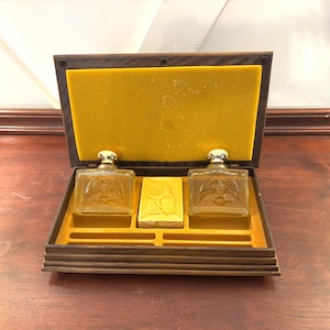 May include: A vintage wooden box with a yellow interior houses two glass bottles with eagle designs and a small gold-colored item. The bottles have silver-colored caps. The box is open, revealing the contents.