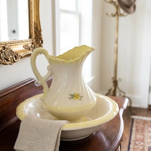 May include: A cream-colored ceramic pitcher and matching washbasin set featuring a scalloped rim and a delicate yellow floral decal. A folded light beige linen napkin with a stitched border rests on the edge of the basin.