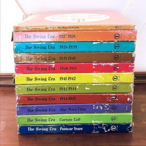 May include: A stack of vintage vinyl record albums titled "The Swing Era" in various colors. Each album spine displays the year range, such as "1937-1938" and "1938-1939". The albums are stacked on a wooden surface.