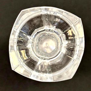 May include: Clear glass decorative bowl with a square shape and a circular interior. The bowl has a textured surface and is designed with a series of radiating lines. The bowl is transparent, allowing light to pass through.