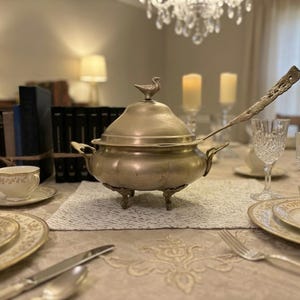 May include: A silver-toned metal tureen with a lid featuring a bird-shaped finial, two side handles, and three ornate feet. A long-handled matching ladle rests inside the bowl. The tureen has a rounded body and a smooth, metallic finish.