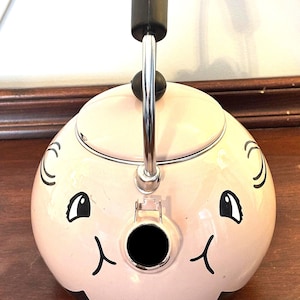 May include: A light pink, round tea kettle with a black handle and spout. The kettle features a whimsical design with black cartoon eyes, a nose, and a mouth. The handle is silver and black.