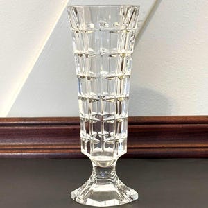 May include: A clear, vintage-style glass vase with a geometric, grid-like pattern. The vase has a flared top and a pedestal base. The vase is approximately 8 inches tall and is designed to hold flowers or other decorative items.