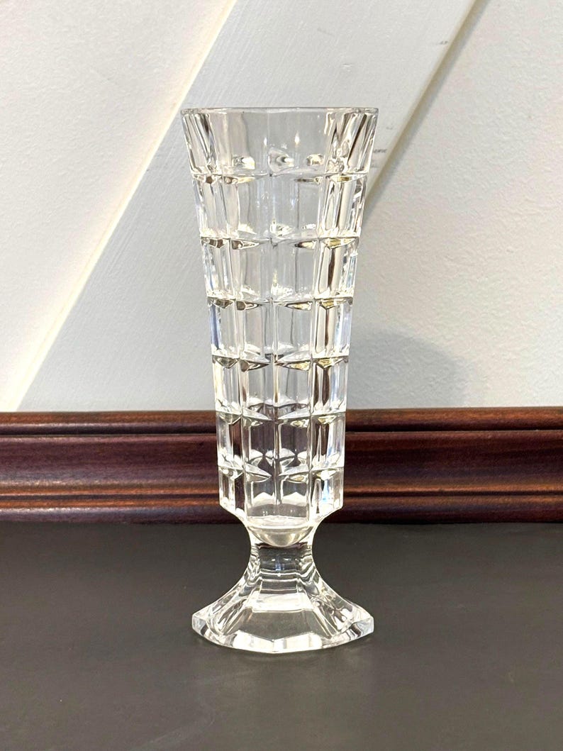 May include: A clear, vintage-style glass vase with a geometric, grid-like pattern. The vase has a flared top and a pedestal base. The vase is approximately 8 inches tall and is designed to hold flowers or other decorative items.